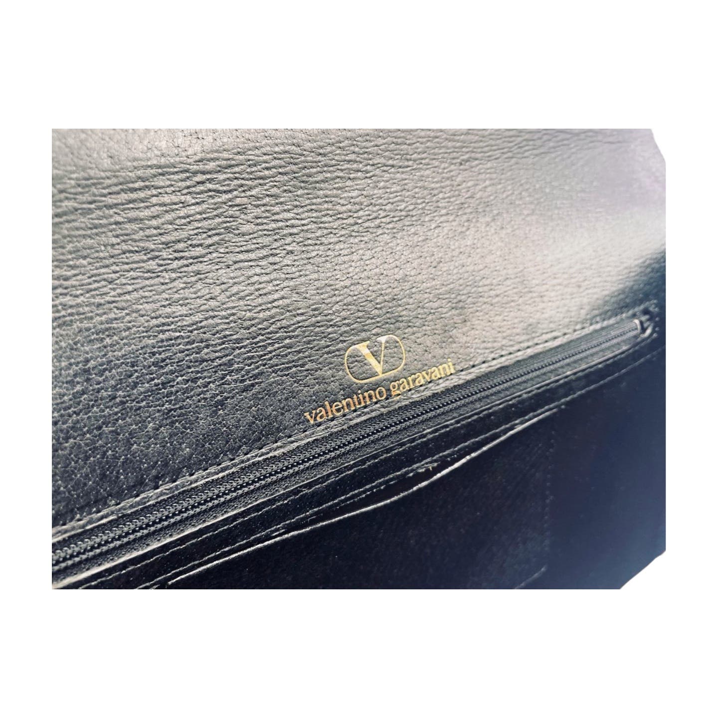 Close-up image of a Valentino Garavani Valentino Black Leather Clutch featuring the "Valentino Garavani" logo embossed in gold above a zippered pocket. The texture of the grained leather is visible.