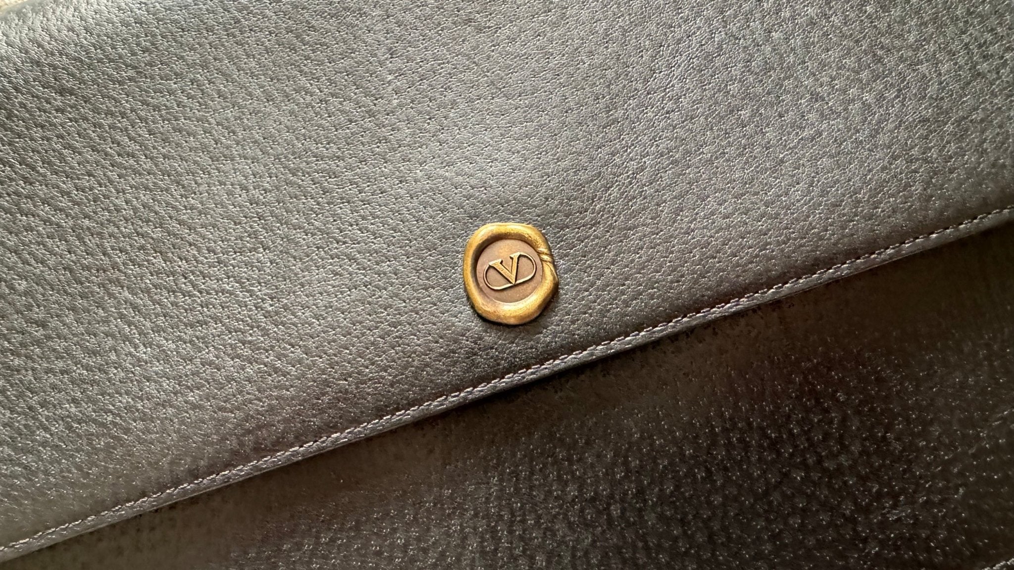 Close-up of a vintage Valentino Garavani Valentino Black Leather Clutch in textured dark gray grained leather, featuring a small, round, gold emblem with interlocked initials. The emblem is centered near the top edge of the bag. The original Valentino dust bag adds an extra touch of elegance.