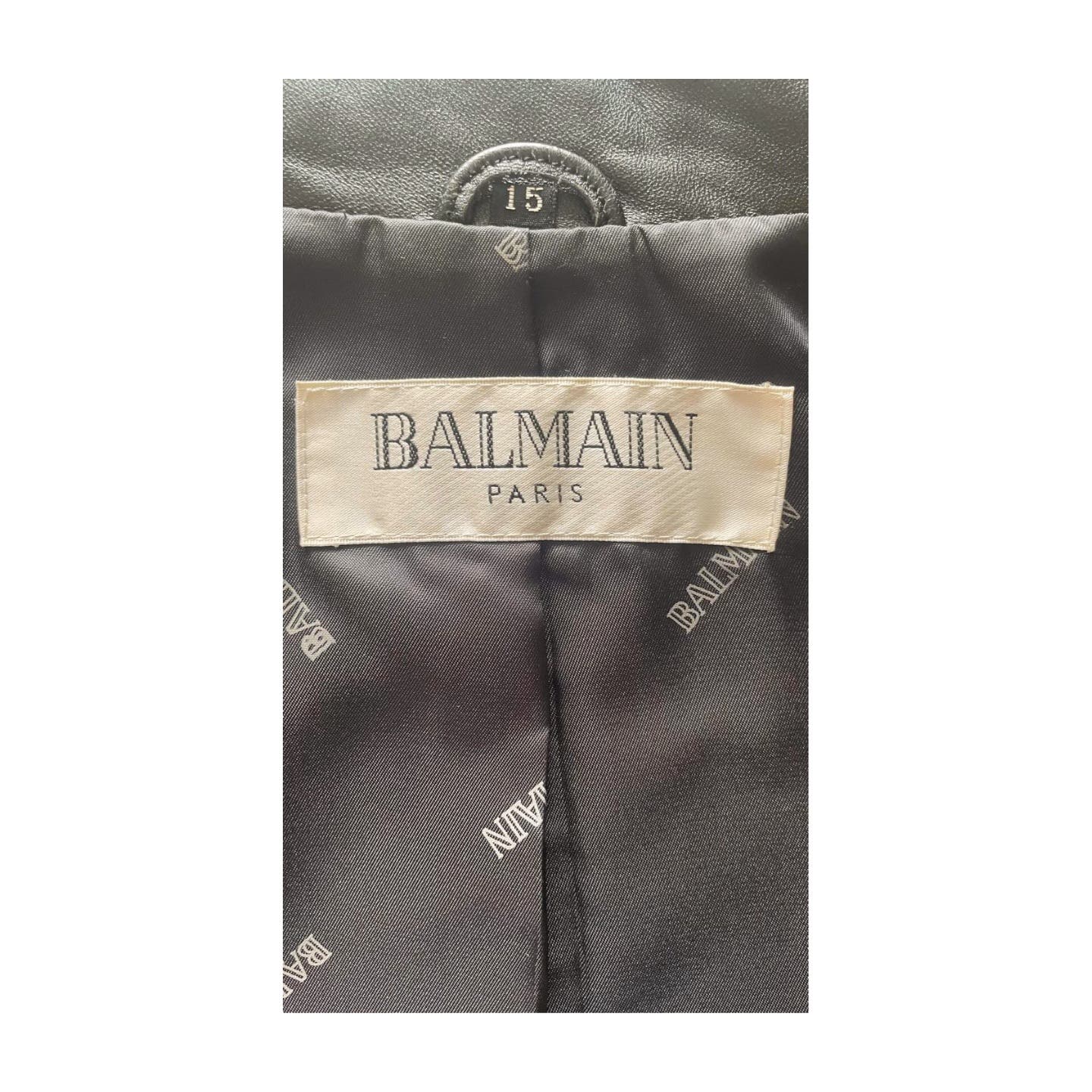 Close-up image of a Balmain label stitched inside a Vintage Balmain Black Leather Blazer. The cream-colored label, printed with "BALMAIN PARIS" in black uppercase letters, sits atop a polyester lining that features a repeated pattern of the word "BALMAIN" in light gray text. Professional cleaning recommended.