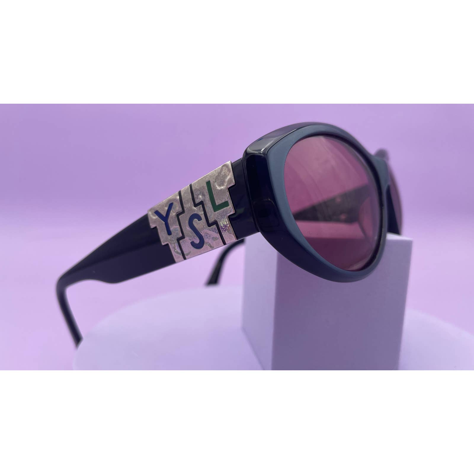 A pair of Saint Laurent YSL Vintage Logo Sunglasses with round, tinted lenses in excellent condition is displayed on a small, white stand against a light purple background. The arms feature intricate silver accents with bold, stylized lettering, exuding a touch of style.