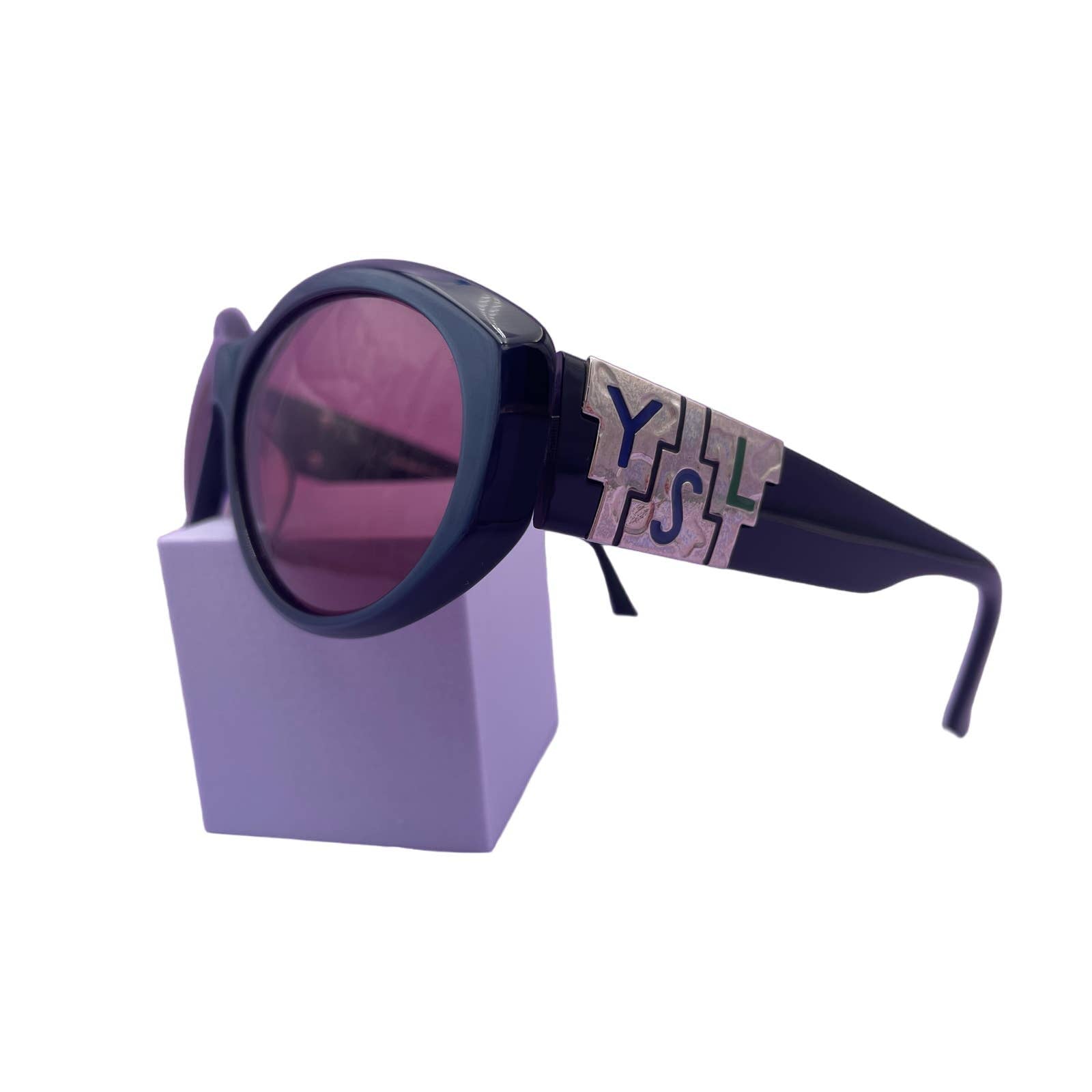 A pair of Saint Laurent YSL Vintage Logo Sunglasses in excellent condition with dark frames and purple-tinted lenses is displayed on a light purple stand. The sunglasses feature a distinctive silver decorative piece with letters, adding a unique touch to the overall style.