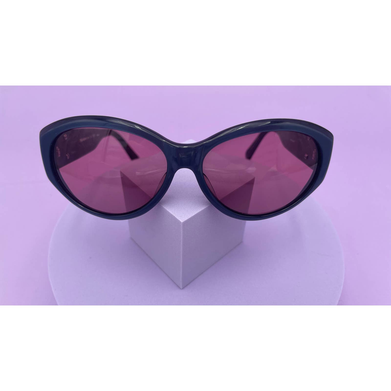 A pair of Saint Laurent YSL Vintage Logo Sunglasses with bold red lenses, in excellent condition, rests on a white display stand against a pastel purple background. The stylish Saint Laurent sunglasses have a classic cat-eye shape, and the display stand features a simple, minimalist design.