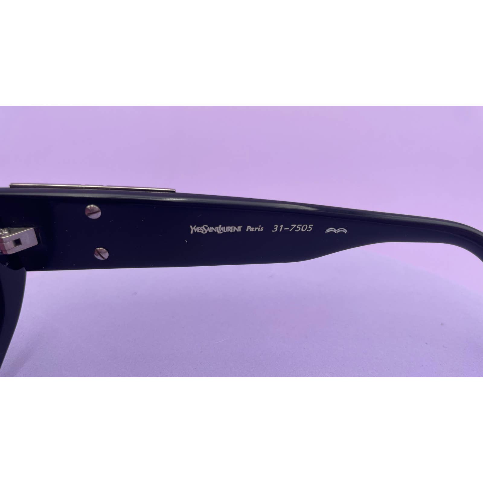 Close-up of the arm of a pair of Saint Laurent YSL Vintage Logo Sunglasses against a lavender background. The arm, in excellent condition, is embossed with the text "Yves Saint Laurent Paris 31-7505" in white lettering and features a small logo, showcasing impeccable style.