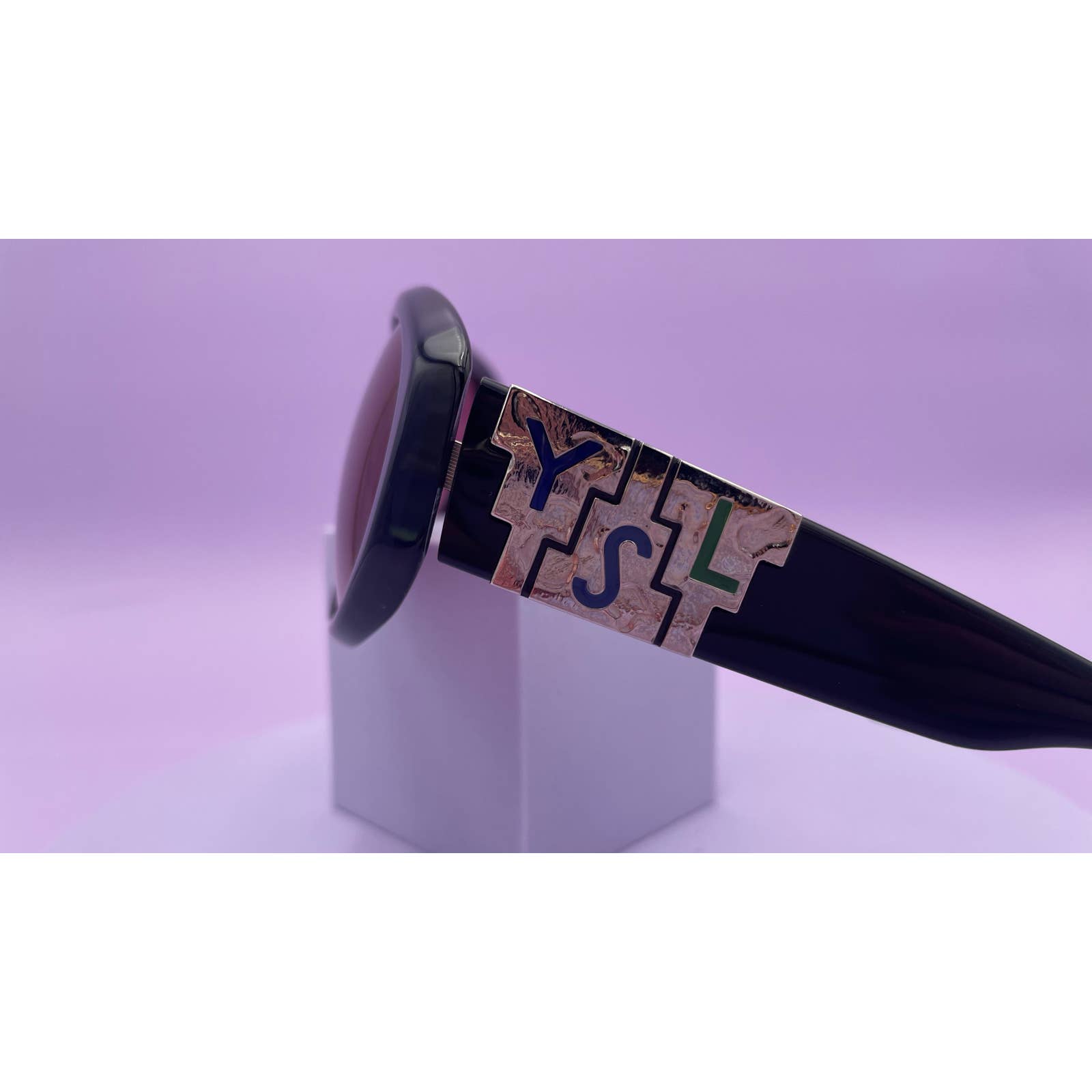 A close-up of a large, dark sunglass frame against a pastel purple background. The focus is on the embossed pattern on the temple, which reads "YSL" in an intricate, stylized design with a marble-like texture. This Saint Laurent YSL Vintage Logo Sunglasses frame exemplifies style and is in excellent condition.