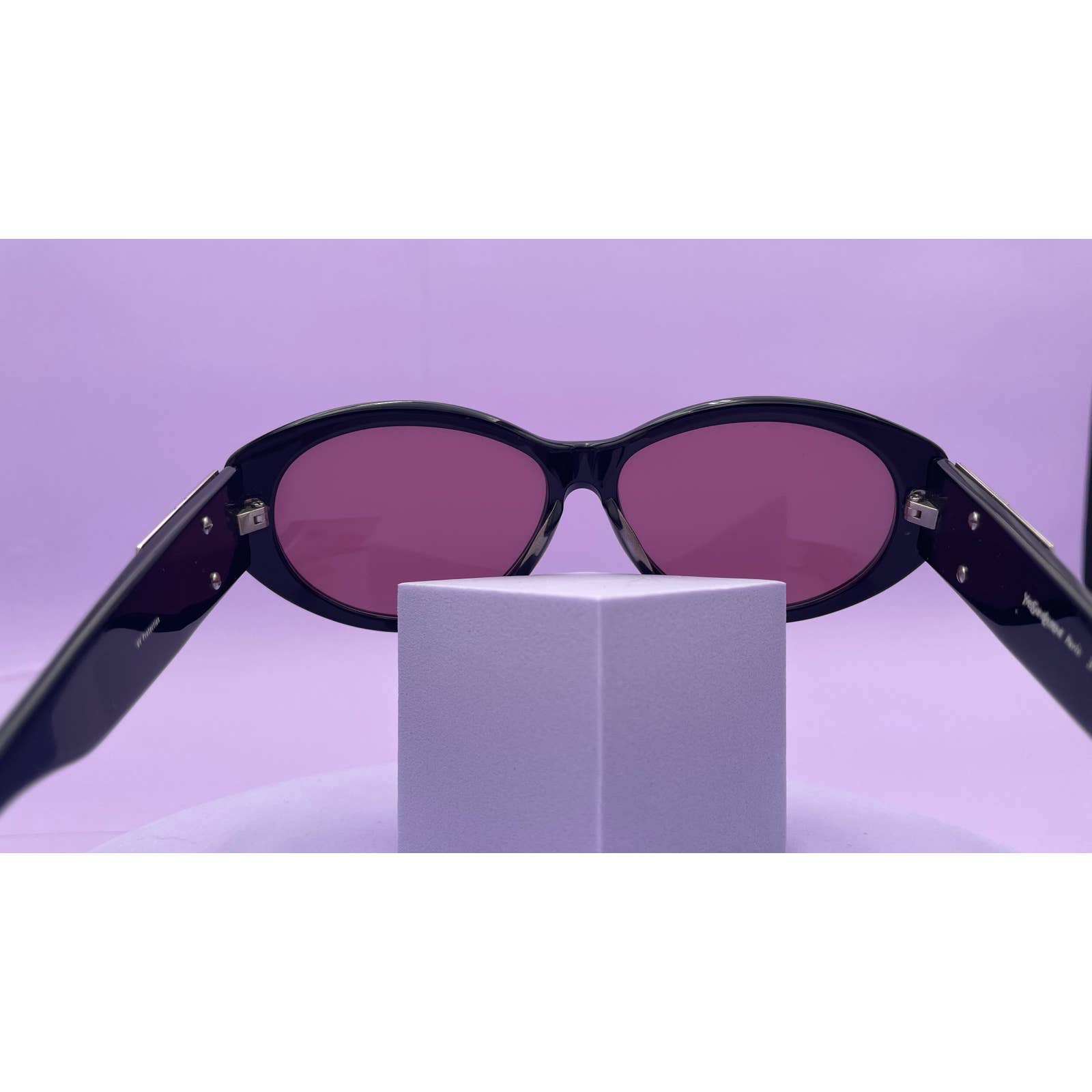 A pair of Saint Laurent YSL Vintage Logo Sunglasses with sleek black frames and purple lenses is open, resting on a small, light purple pedestal. The background is also light purple, creating a stylish monochromatic look. These sunglasses are in excellent condition.