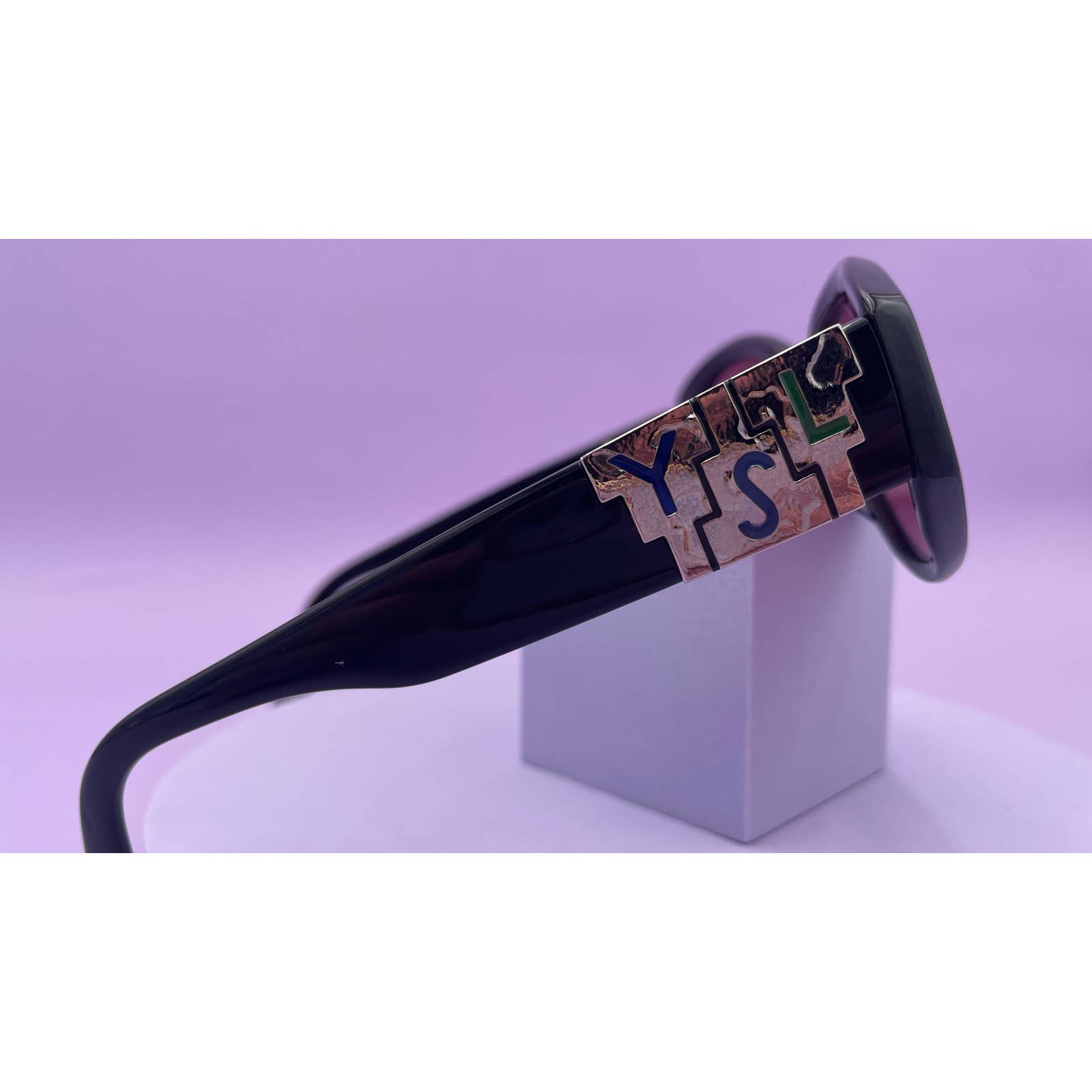 Close-up of the black arm of a pair of Saint Laurent YSL Vintage Logo Sunglasses in excellent condition, featuring a stylized design with the acronym "YSL" in gold on the hinge. The glasses rest on a small white box with a purple background, showcasing their impeccable style.