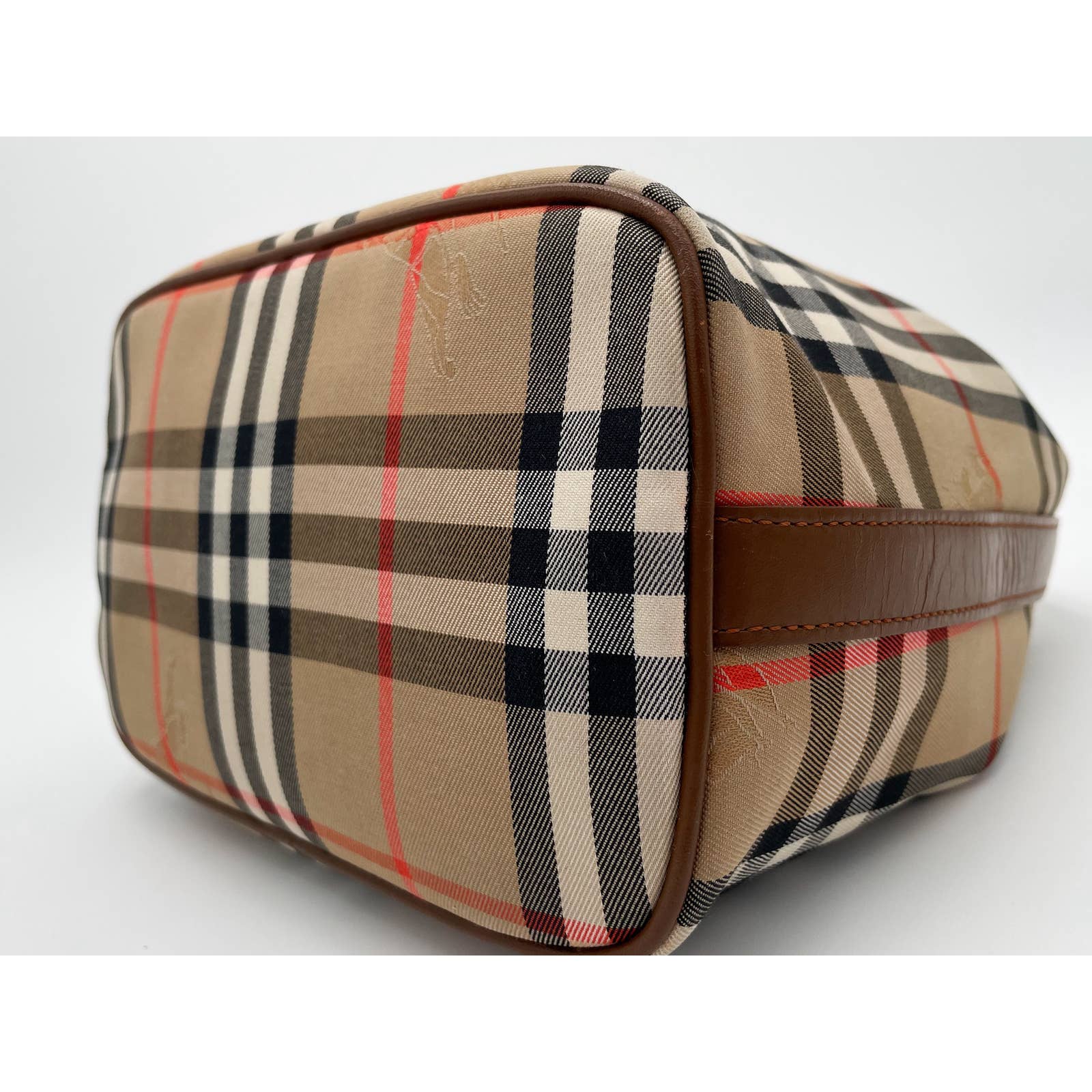 A rectangular, Burberry Vintage House Check Bag with a classic beige, black, and red plaid design is shown. The canvas bag features a zip top and brown leather trim on its side, set against a white background. The image focuses on the bottom and one side of the bag.